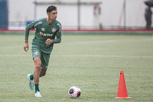 Fotografia de ação do jogador Vitor Mendes durante um treinamento em campo gramado sob chuva leve. Ele está em movimento de corrida, conduzindo uma bola de futebol oficial. Veste um conjunto de treino na cor verde escuro com detalhes brancos e o escudo de um clube anterior. Ao lado dele, vê-se um cone de sinalização laranja no gramado.