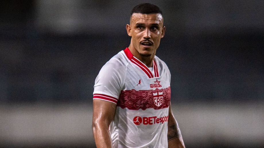 "Fotografia colorida do jogador William Pottker vestindo o uniforme branco do CRB. Ele é um homem pardo, com cabelo curto cortado em degradê e um bigode fino. O atleta aparece suado, com expressão focada, olhando para o lado durante uma partida noturna. A camisa possui detalhes em vermelho na gola e nas mangas, com o escudo do clube e o patrocínio 'BETesporte' em destaque."