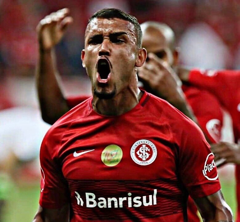 "Close do jogador William Pottker em um momento de intensa celebração. Ele está com a boca aberta, gritando de emoção, com os músculos do pescoço tensionados. Veste a camisa vermelha do Internacional, com o escudo do clube e o patrocínio da Banrisul visíveis. Ao fundo, outros jogadores e a torcida aparecem desfocados, ressaltando a vibração do atleta."