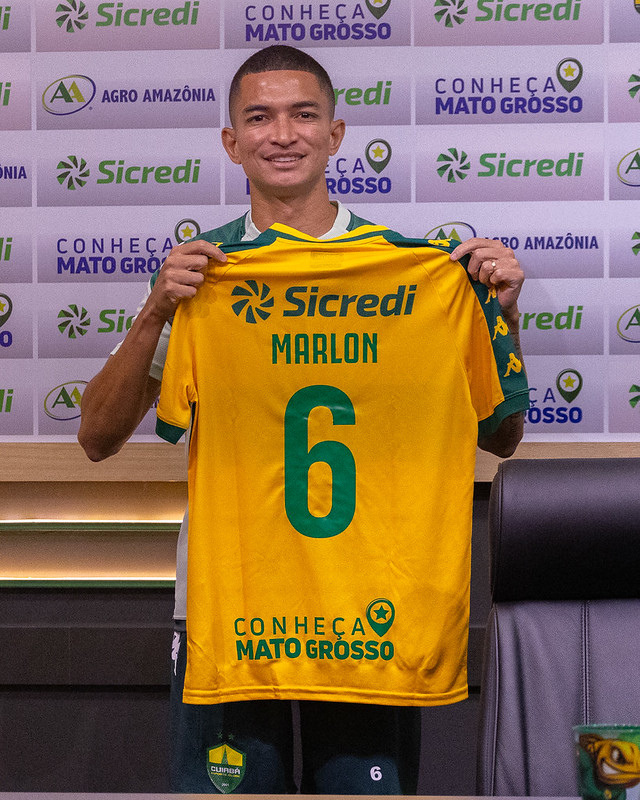 Lateral Marlon sorri para a câmera enquanto segura a camisa oficial de futebol do Cuiabá Esporte Clube. A camisa é amarela com detalhes verdes, exibindo o nome "MARLON" e o número 6. Ao fundo, um painel publicitário com os logotipos "Sicredi", "Agro Amazônia" e a frase "Conheça Mato Grosso".
