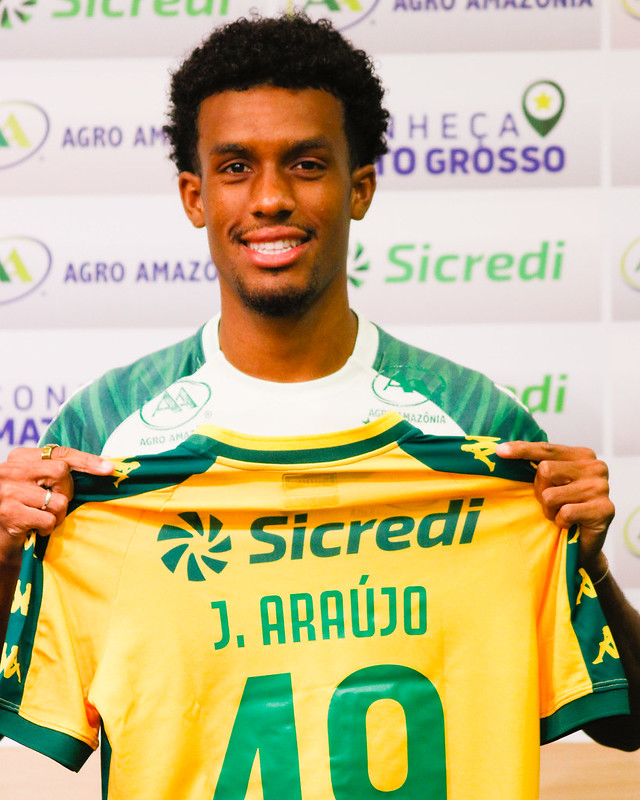 O jogador J. Araújo sorri para a foto segurando a camisa amarela de jogo do Cuiabá. A camisa destaca o nome "J. ARAÚJO" e o número 40. Ele usa anéis na mão esquerda. O cenário de fundo é o mesmo painel de patrocinadores das imagens anteriores.