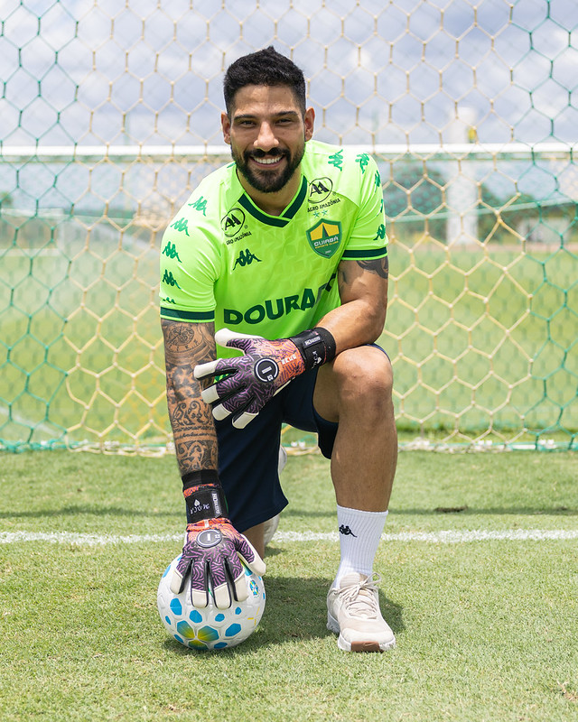 Marcelo Carné sorri ajoelhado no campo do Cuiabá, segurando a bola e vestindo uniforme verde do Dourado.