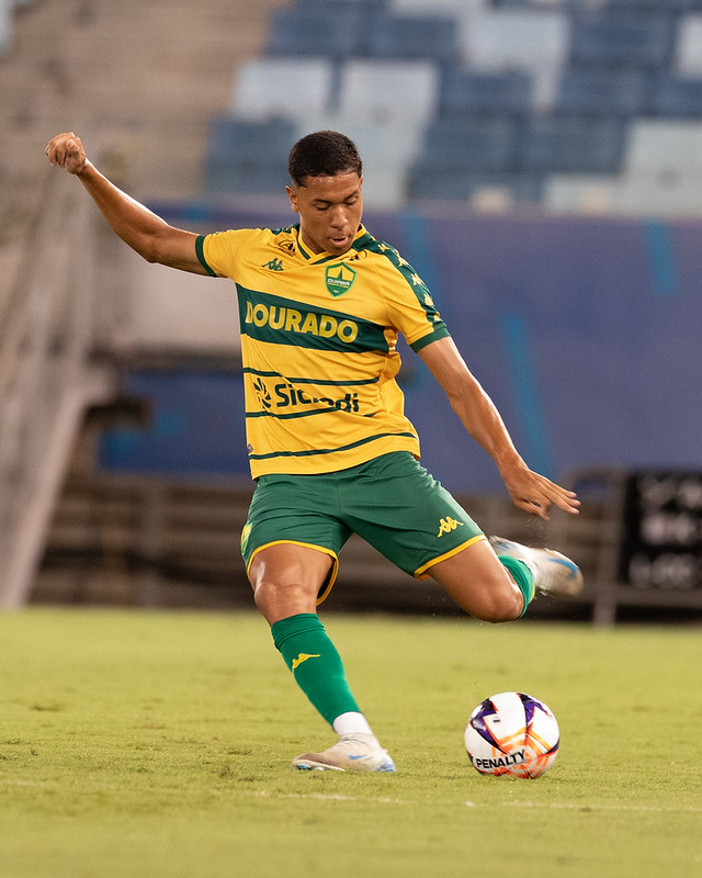 Fotografia em plano médio de um jogador do Cuiabá, jovem e de pele parda, em ação no campo. Ele veste o uniforme oficial amarelo com listras verdes horizontais e calções verdes. O atleta está inclinado para a direita, com a perna esquerda de apoio e a direita esticada após chutar uma bola de futebol da marca Penalty. O fundo mostra as cadeiras desfocadas das arquibancadas da Arena Pantanal durante a noite.