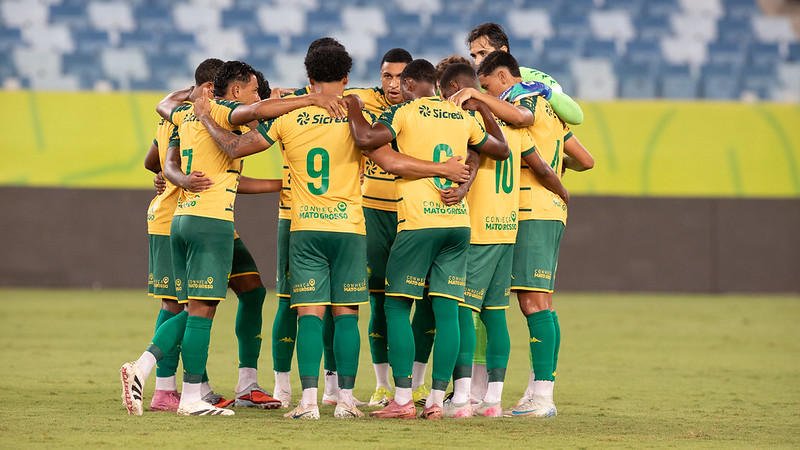 Foto do elenco do Cuiabá reunido em um círculo no centro do gramado. Os jogadores estão de costas, abraçados pelos ombros em um momento de união antes ou depois da partida. É possível ver os números 7, 9, 8 e 10 nos uniformes amarelos, além do goleiro à direita vestindo uniforme verde claro. Ao fundo, as arquibancadas do estádio estão vazias e iluminadas pelos refletores.