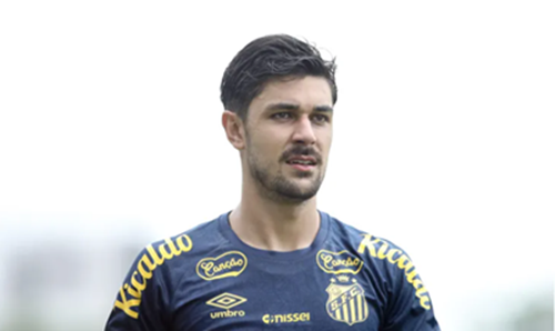 Retrato de meio corpo do zagueiro João Basso durante um treino ou aquecimento. Ele está posicionado levemente de perfil, com uma expressão séria e focada, olhando para o lado. Veste uma camisa de treino azul marinho do Santos FC, com detalhes em amarelo e o escudo do clube no peito. O fundo é neutro e claro, mantendo o foco total no atleta.