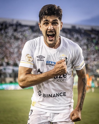 Fotografia em plano médio do zagueiro João Basso, do Santos FC, comemorando um gol. Ele está de frente, com a boca aberta em um grito de celebração e os punhos cerrados à frente do peito. Veste o uniforme branco oficial com detalhes dourados e diversos patrocinadores. Ao fundo, a arquibancada do estádio está lotada de torcedores, criando um efeito levemente desfocado que destaca a expressão de euforia do atleta.
