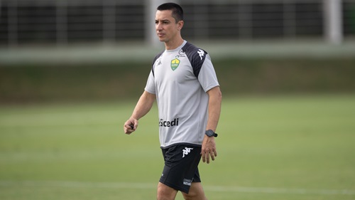 Fotografia em plano médio do técnico Eduardo Barros durante um treinamento no Cuiabá Esporte Clube. Ele é um homem de pele clara, cabelos curtos e escuros, e veste o uniforme de treino oficial: uma camiseta cinza com detalhes pretos nos ombros, o escudo do clube no peito e o patrocínio da Sicredi. Ele usa um relógio de pulso preto e caminha pelo gramado verde, que aparece levemente desfocado ao fundo.