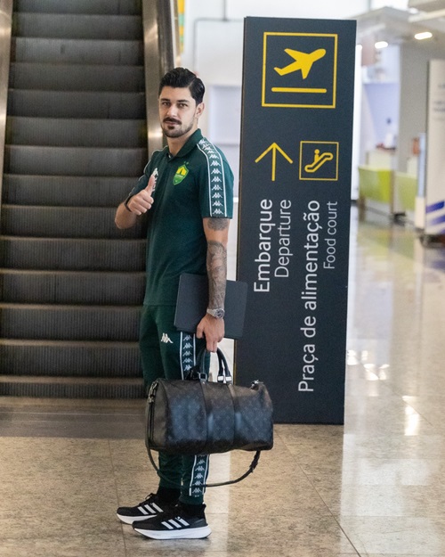 Fotografia de corpo inteiro de um jogador do Cuiabá posando em um aeroporto. O atleta está de pé ao lado de uma placa de sinalização de embarque e praça de alimentação. Ele veste o conjunto de viagem oficial do clube na cor verde escuro com detalhes brancos da marca Kappa. O jogador segura uma pasta preta e uma mala de mão escura, fazendo um sinal de "joinha" com a mão direita e sorrindo para a câmera. Ao fundo, vê-se uma escada rolante.