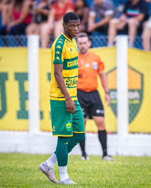 Gabriel Mineiro do Cuiabá em campo durante partida, observando o jogo, com uniforme verde e amarelo e arbitragem ao fundo.