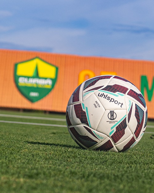 Bola oficial de futebol posicionada sobre o gramado, com o escudo do Cuiabá ao fundo em um painel laranja desfocado.