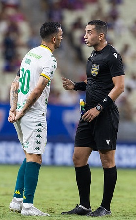 Jogador do Cuiabá discute com árbitro durante partida, em campo, demonstrando reclamação em lance do jogo.