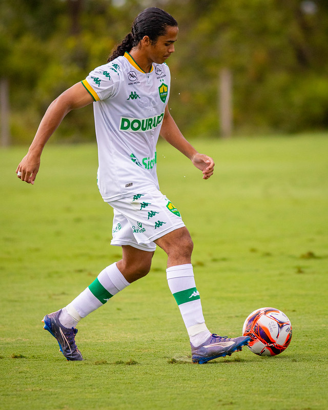 Jogador do Cuiabá, vestindo uniforme branco com detalhes verdes, dominando a bola com o pé direito em um campo gramado, com foco e concentração durante a atividade.