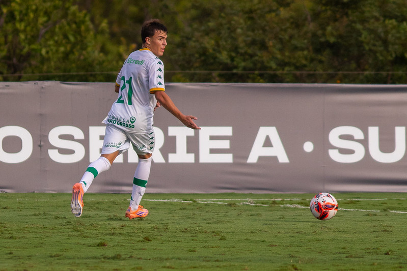 Jogador do Cuiabá Sub-20, vestindo uniforme branco com detalhes em verde e número 21, correndo próximo à linha lateral enquanto acompanha a bola em campo gramado.