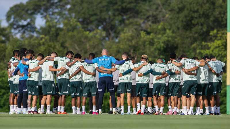 O elenco e a comissão técnica do Cuiabá formam um grande círculo (roda) no campo de treinamento. Os jogadores estão de costas, abraçados, enquanto um integrante da equipe técnica vestindo azul está no centro. O cenário mostra um gramado bem cuidado com árvores ao fundo sob a luz do dia.