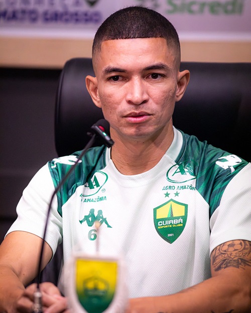 Lateral Marlon concede entrevista coletiva pelo Cuiabá, sentado à mesa com microfone e escudo do clube ao fundo.