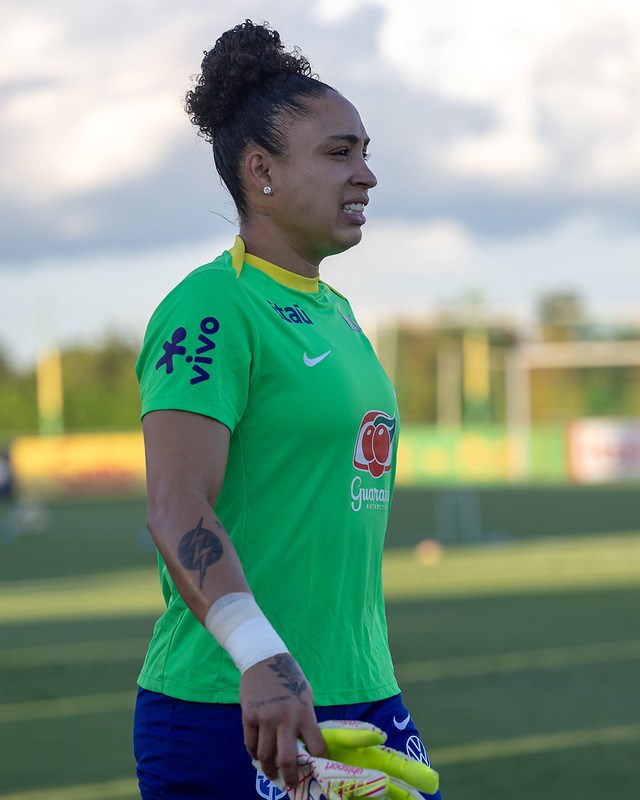 Close da Goleira da seleção brasileira feminina com uma camisa de treino verde vibrante. Ela está segurando suas luvas e parece concentrada ou em meio a uma conversa durante uma pausa no treino.