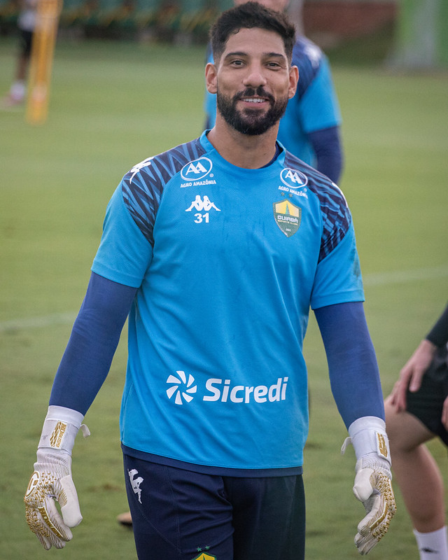 Goleiro do Cuiabá Esporte Clube, Marcelo Carné, sorrindo durante treino no campo, vestindo uniforme azul com luvas, em preparação para jogos da Série B.