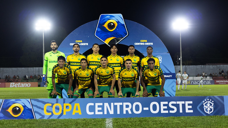 Time titular do Cuiabá posando para foto oficial antes da partida da Copa Centro-Oeste, com placa da competição ao fundo e estádio iluminado à noite.