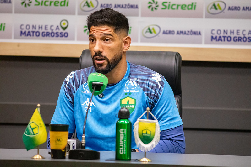 Marcelo Carné durante entrevista coletiva do Cuiabá Esporte Clube, falando ao microfone em sala de imprensa, com backdrop de patrocinadores ao fundo.
