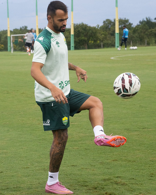 Um jogador de futebol posa para a foto enquanto equilibra uma bola de futebol no joelho esquerdo (embaixadinha) em um campo de treino. Ele veste uma camisa de treino verde-clara com detalhes em verde-escuro nos ombros, shorts verde-escuro com o escudo do Cuiabá Esporte Clube e chuteiras de cor rosa vibrante. O jogador tem tatuagens na perna direita e olha atentamente para a bola. O fundo mostra um campo gramado amplo com outros jogadores e postes de iluminação distantes.