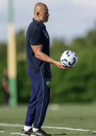 Treinador do Cuiabá em campo de treino segurando uma bola, vestindo uniforme azul do clube, com o gramado e fundo desfocado.
