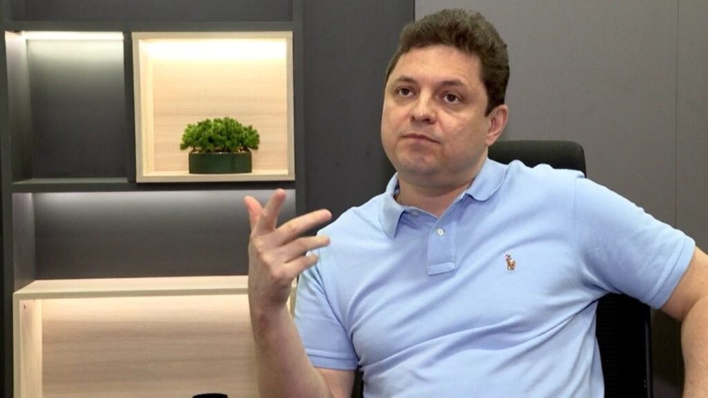 Cristiano dresch sentado em uma cadeira durante entrevista, vestindo camisa polo azul clara, gesticulando com a mão enquanto fala, com expressão séria e ambiente moderno ao fundo.