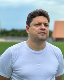 Cristiano Dresch em pé ao ar livre, vestindo camiseta clara, com expressão concentrada, tendo um campo e estruturas ao fundo em ambiente aberto.