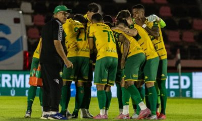Um grupo de jogadores de futebol com uniformes amarelos e verdes, e um treinador, se reúnem em campo, possivelmente durante uma discussão tática ou celebração. O treinador, um homem mais velho com óculos e boné, está à esquerda, olhando para os jogadores que estão aglomerados no centro.