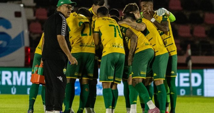 Um grupo de jogadores de futebol com uniformes amarelos e verdes, e um treinador, se reúnem em campo, possivelmente durante uma discussão tática ou celebração. O treinador, um homem mais velho com óculos e boné, está à esquerda, olhando para os jogadores que estão aglomerados no centro.