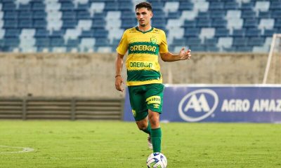O jogador Renan Carrasco, em campo, vestindo o uniforme amarelo e verde do Cuiabá. Ele está conduzindo a bola com o pé direito em um estádio vazio.
