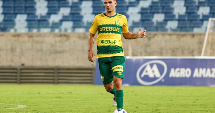 O jogador Renan Carrasco, em campo, vestindo o uniforme amarelo e verde do Cuiabá. Ele está conduzindo a bola com o pé direito em um estádio vazio.