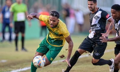 Fotografia de uma partida de futebol em campo de grama natural. Em primeiro plano, um jogador do Cuiabá, vestindo camisa amarela e calções verdes, está em movimento dinâmico tentando controlar a bola com o corpo inclinado. Ele é marcado de perto por dois jogadores adversários que vestem uniformes pretos com detalhes brancos. A ação levanta um pouco de terra do gramado, e o fundo mostra outros jogadores e torcedores de forma desfocada.