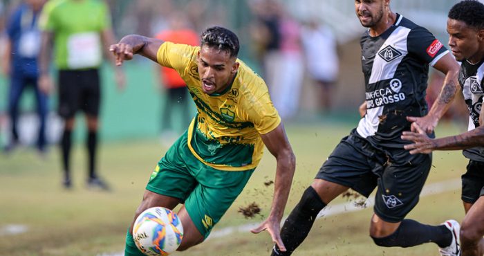 Fotografia de uma partida de futebol em campo de grama natural. Em primeiro plano, um jogador do Cuiabá, vestindo camisa amarela e calções verdes, está em movimento dinâmico tentando controlar a bola com o corpo inclinado. Ele é marcado de perto por dois jogadores adversários que vestem uniformes pretos com detalhes brancos. A ação levanta um pouco de terra do gramado, e o fundo mostra outros jogadores e torcedores de forma desfocada.