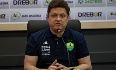 presidente Cristiano Dresch do Cuiabá fala em coletiva de imprensa, sentado à mesa com microfone, usando camisa polo do clube e painel com patrocinadores ao fundo.