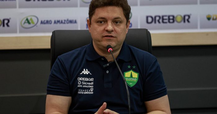 presidente Cristiano Dresch do Cuiabá fala em coletiva de imprensa, sentado à mesa com microfone, usando camisa polo do clube e painel com patrocinadores ao fundo.