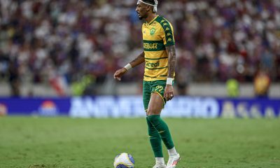 jogador Derik do Cuiabá Esporte Clube durante uma partida. Ele é um homem negro com o cabelo preso em tranças curtas e loiras, vestindo o uniforme titular amarelo com listras verdes horizontais, calções verdes e meiões verdes. Ele está parado no gramado com o pé esquerdo próximo à bola, olhando atentamente para o lado. O fundo mostra as arquibancadas de um estádio lotado e desfocado.