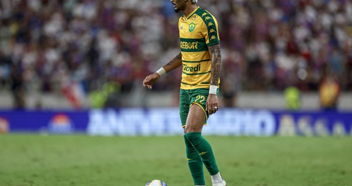 jogador Derik do Cuiabá Esporte Clube durante uma partida. Ele é um homem negro com o cabelo preso em tranças curtas e loiras, vestindo o uniforme titular amarelo com listras verdes horizontais, calções verdes e meiões verdes. Ele está parado no gramado com o pé esquerdo próximo à bola, olhando atentamente para o lado. O fundo mostra as arquibancadas de um estádio lotado e desfocado.