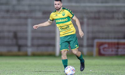 "Fotografia colorida do jogador Léo Ataíde, do Cuiabá, durante uma partida de futebol. Ele é um homem pardo, de barba aparada e cabelo curto, vestindo o uniforme principal do clube: camisa amarela com detalhes verdes e a inscrição 'DOURADO', calções verdes e meiões verdes. Ele está em movimento no gramado, conduzindo a bola com o pé direito. O fundo mostra as arquibancadas de um estádio de forma desfocada."
