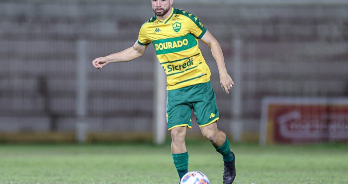 "Fotografia colorida do jogador Léo Ataíde, do Cuiabá, durante uma partida de futebol. Ele é um homem pardo, de barba aparada e cabelo curto, vestindo o uniforme principal do clube: camisa amarela com detalhes verdes e a inscrição 'DOURADO', calções verdes e meiões verdes. Ele está em movimento no gramado, conduzindo a bola com o pé direito. O fundo mostra as arquibancadas de um estádio de forma desfocada."
