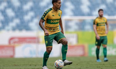 O volante Denilson em campo, visto de corpo inteiro em um ângulo lateral. Ele utiliza chuteiras brancas e controla uma bola de futebol oficial sobre o gramado. O uniforme da marca Kappa destaca o patrocínio master "Dourado" e o selo da "Sicredi". Outro jogador do Cuiabá aparece ao fundo, fora de foco, observando a jogada.