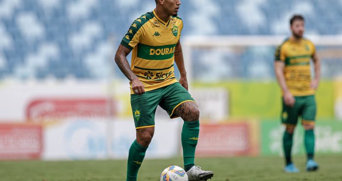 O volante Denilson em campo, visto de corpo inteiro em um ângulo lateral. Ele utiliza chuteiras brancas e controla uma bola de futebol oficial sobre o gramado. O uniforme da marca Kappa destaca o patrocínio master "Dourado" e o selo da "Sicredi". Outro jogador do Cuiabá aparece ao fundo, fora de foco, observando a jogada.