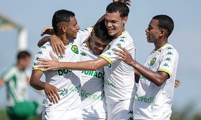 Jogadores do Cuiabá Sub-20 comemoram gol com abraços e sorrisos durante partida no CT Manoel Dresch, vestindo uniforme branco com detalhes verdes.