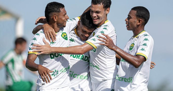 Jogadores do Cuiabá Sub-20 comemoram gol com abraços e sorrisos durante partida no CT Manoel Dresch, vestindo uniforme branco com detalhes verdes.