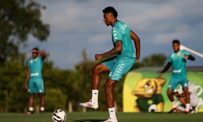 Guilherme Mariano durante treino do Cuiabá, com uniforme verde, controlando a bola no gramado sob o sol, enquanto outros jogadores aparecem ao fundo no centro de treinamento.