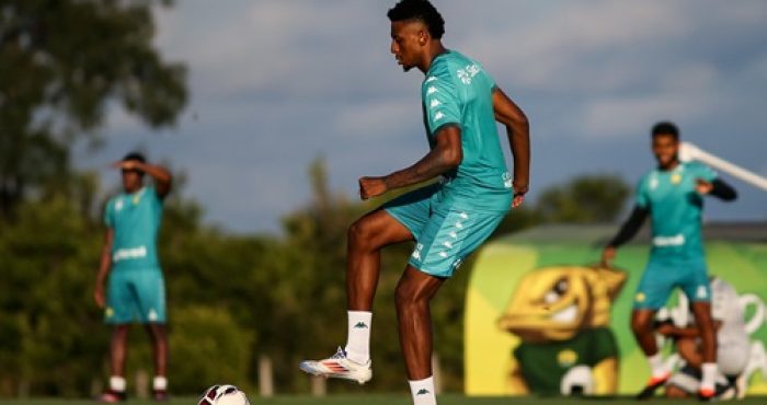 Guilherme Mariano durante treino do Cuiabá, com uniforme verde, controlando a bola no gramado sob o sol, enquanto outros jogadores aparecem ao fundo no centro de treinamento.