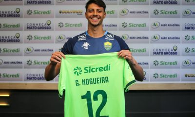 O goleiro Guilherme Nogueira sorrindo e segurando uma camisa verde-clara com o nome 'G. NOGUEIRA' e o número '12' em verde escuro, sobre um fundo com logotipos de patrocinadores.