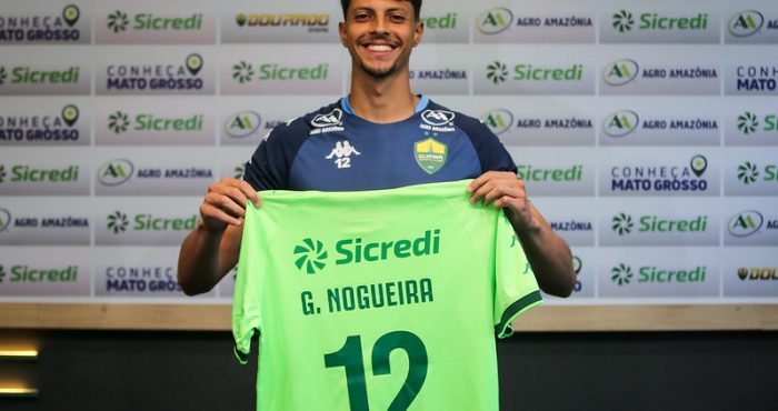 O goleiro Guilherme Nogueira sorrindo e segurando uma camisa verde-clara com o nome 'G. NOGUEIRA' e o número '12' em verde escuro, sobre um fundo com logotipos de patrocinadores.