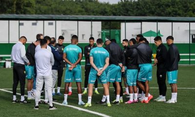 "Grupo de jogadores e comissão técnica do Cuiabá reunidos no centro do campo durante treino, todos usando uniformes de treino verde e preto, com cercas e árvores ao fundo."