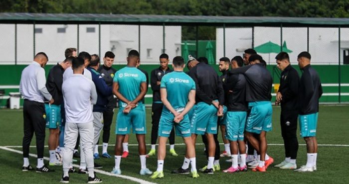 "Grupo de jogadores e comissão técnica do Cuiabá reunidos no centro do campo durante treino, todos usando uniformes de treino verde e preto, com cercas e árvores ao fundo."