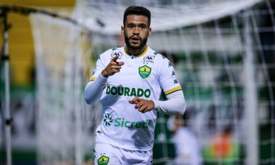 "Patrick de Lucca, volante do Cuiabá, celebra após marcar, apontando com a mão direita. Ele usa o uniforme de jogo branco, em frente a uma trave com rede no fundo."